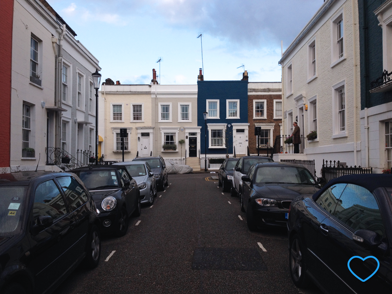 Foto de uma rua em Notting Hill. Casinhas coloridas, mas em tons terrosos. Um senhor na porta de casa abrindo o guarda-chuva.