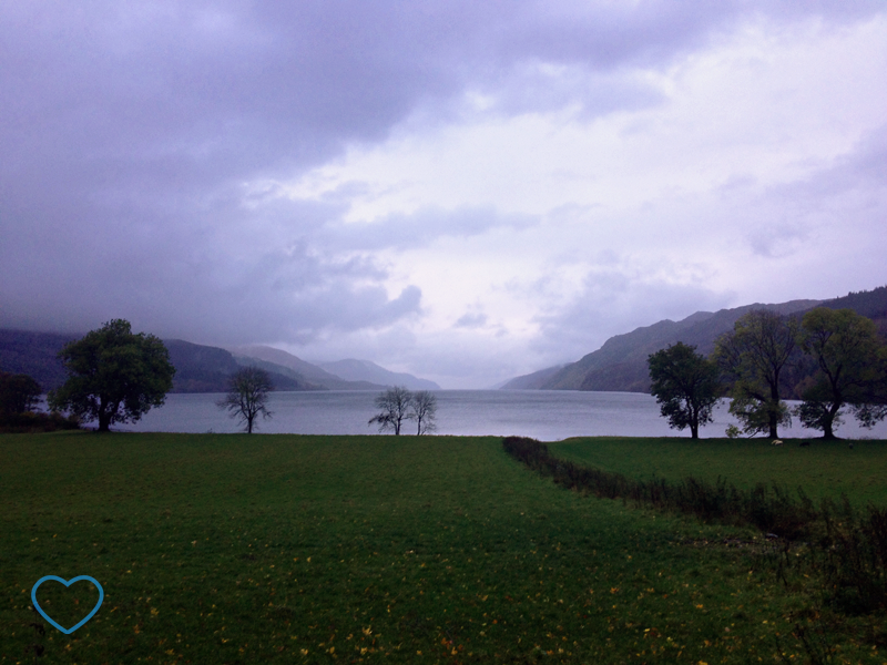 Foto do Lago Ness em um dia nublado. Árvores em volta e gramado.