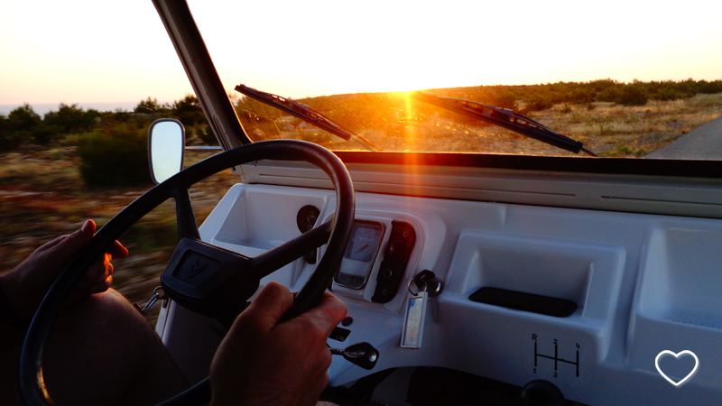 Detalhe de um volante e as mãos de uma pessoa dirigindo um carro sob o sol poente.