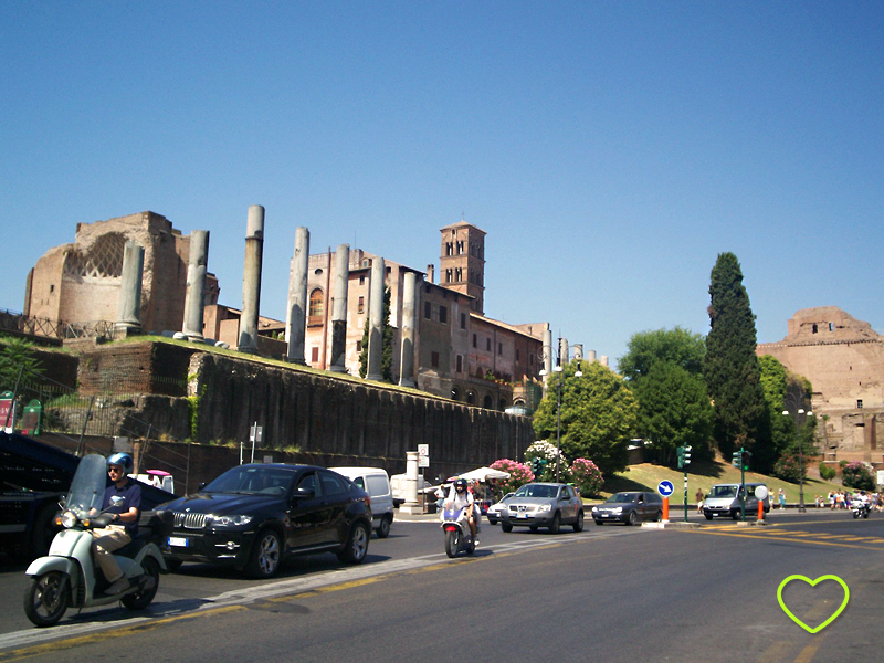 Uma rua em primeiro plano, com automóveis. E em segundo plano, uma construção bem antiga. Acho que é o Foro Romano.