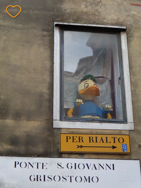Uma janela com um Pato Donald feito de peças de Lego. Abaixo, há uma placa com uma indicação: "Per Rialto".