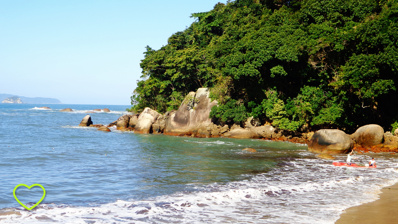 O mar azul em um dia de sol. A espuma que chega do mar à areia é branca e limpa. Uma parte de um monte está como que lançado ao mar e revestido de uma vibrante e verde vegetação.