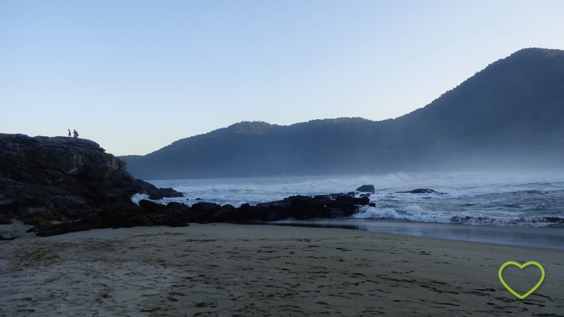 Praia já ao anoitecer. Muitas pedras perto do mar e duas pessoas no alto de uma das pedras.