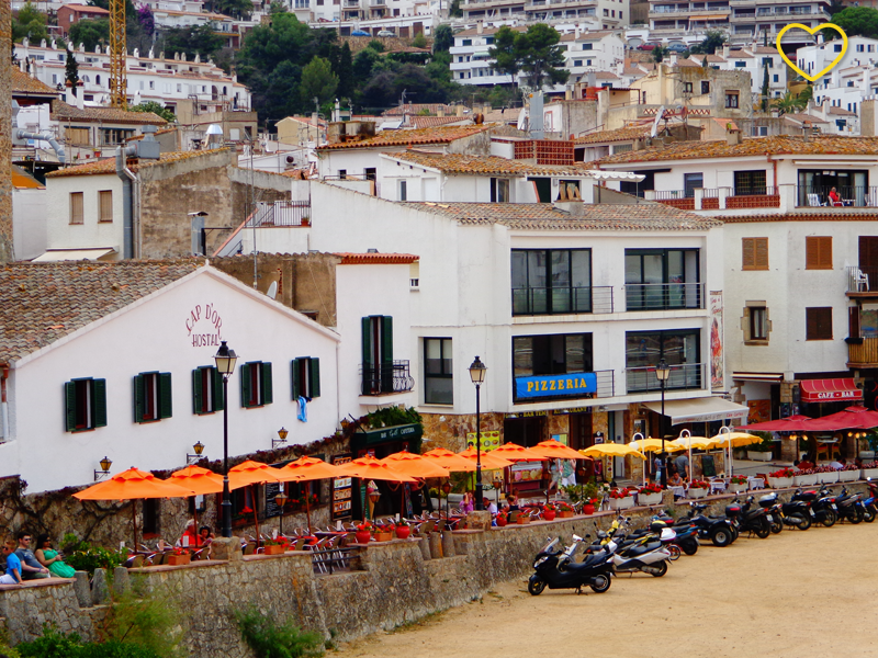 A orla da praia vista da praia. Muitos bares, casas, prédios baixos e mesas com guarda-sóis. E motos estacionadas.