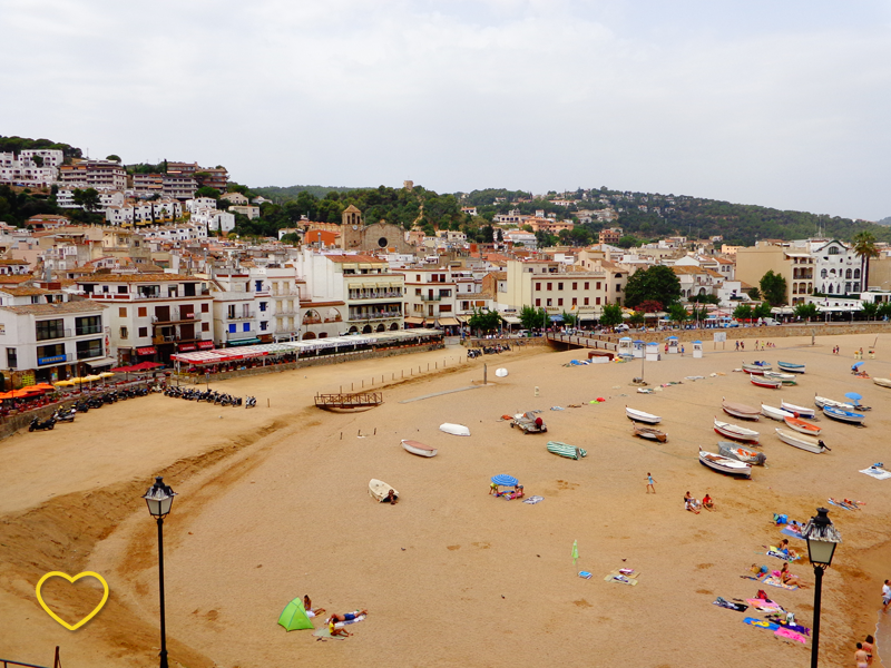 A praia vista do alto do morro. Mas só a areia e a orla estão enquadrados. Muitos barquinhos na areia e muitas casinhas na orla.